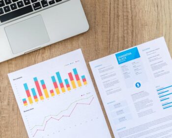 Top view of a laptop, charts, and resume on a wooden desk, showcasing business analysis and job application.